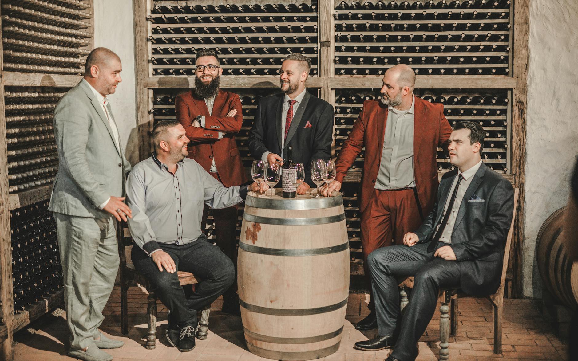 Group of adults in suits enjoying wine in a cozy cellar setting with oak barrels and wine racks.